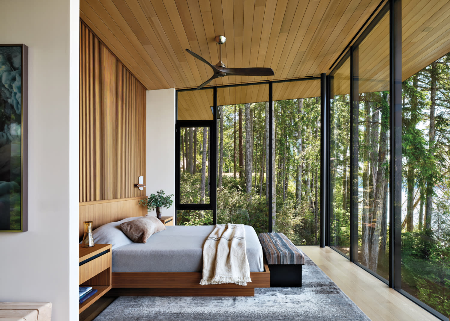 Modern bedroom with wood accents and large glass windows offering a serene forest view. The cozy bed has grey linens and a soft blanket.