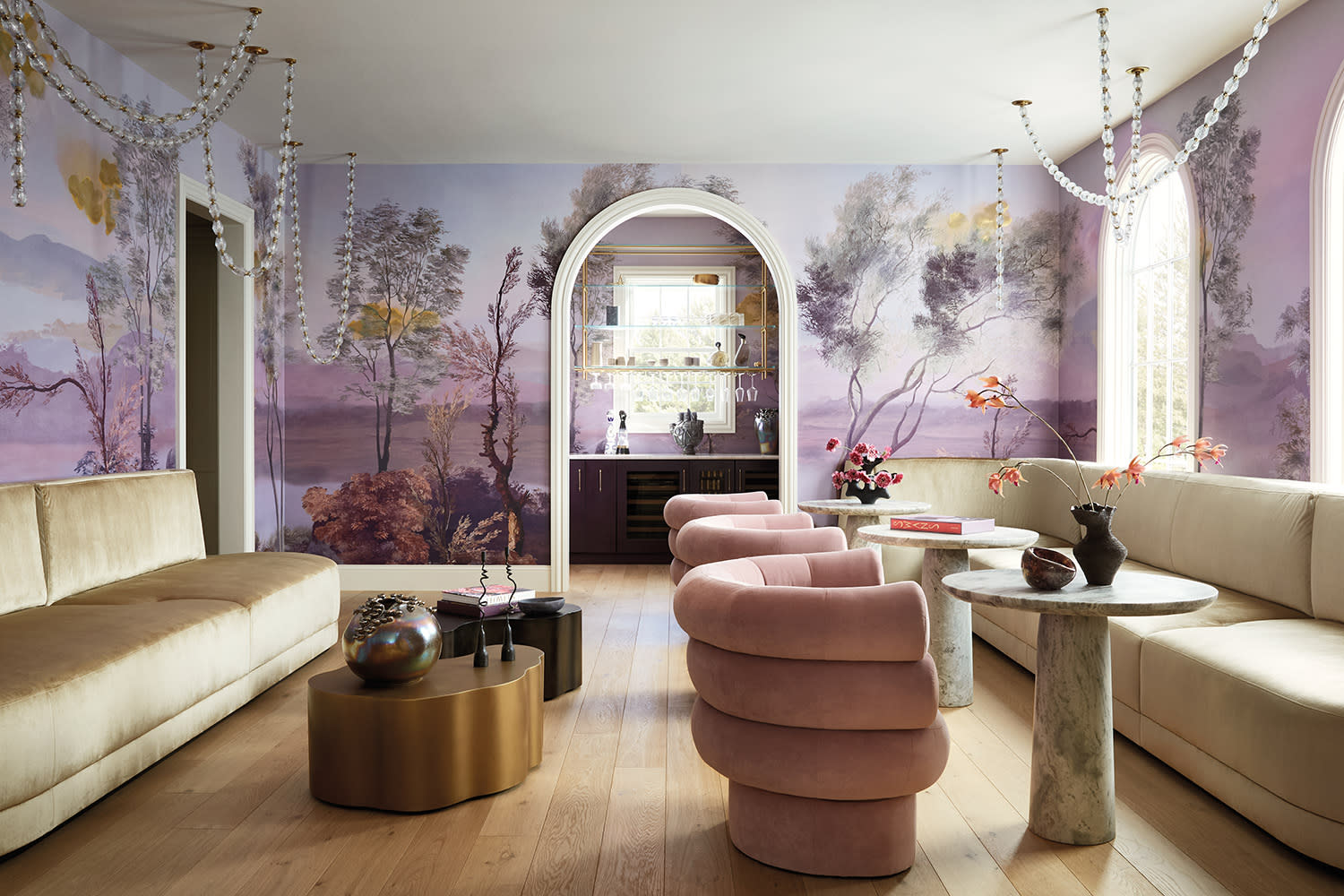 Living room with lavender scenic mural walls, pink curved chairs, beige sofas, and brass-glass chandeliers.