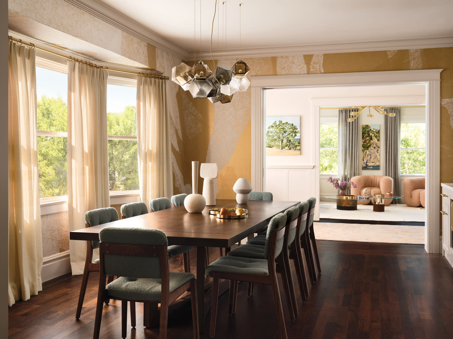 Sophisticated dining room with a dark wooden table and cushioned chairs. Modern pendant lights hang above. Sunlit living room visible through doorway.