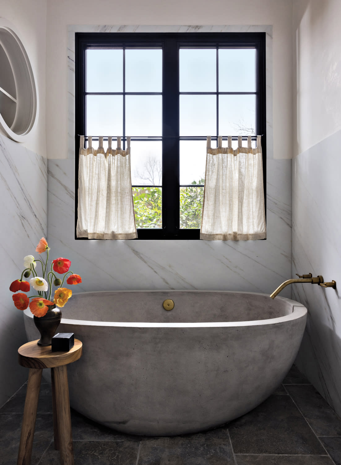 A bathroom with a freestanding concrete tub and brass faucet, sitting under a window with curtains.