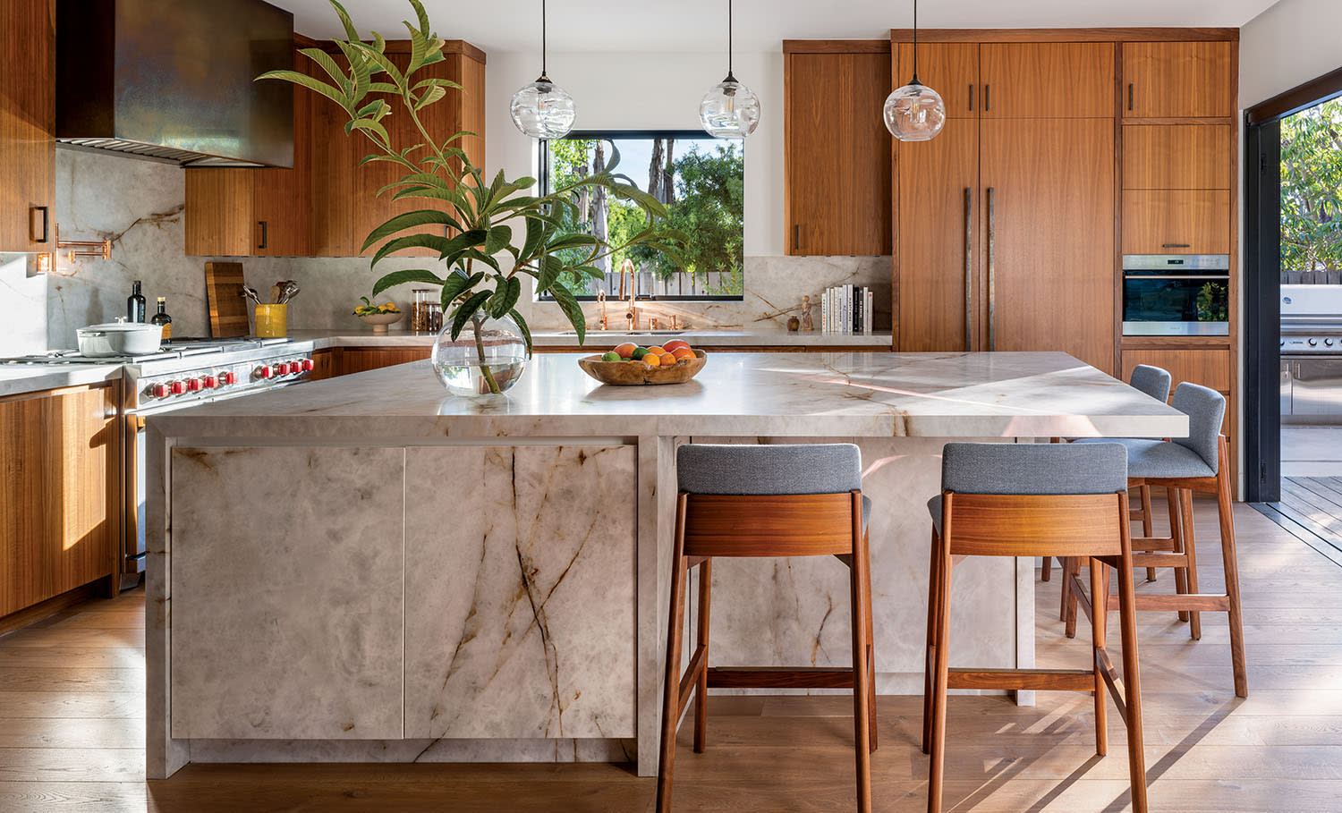 Modern kitchen with warm wood cabinets, marble island, and bar stools. Glass pendant lights hang above.