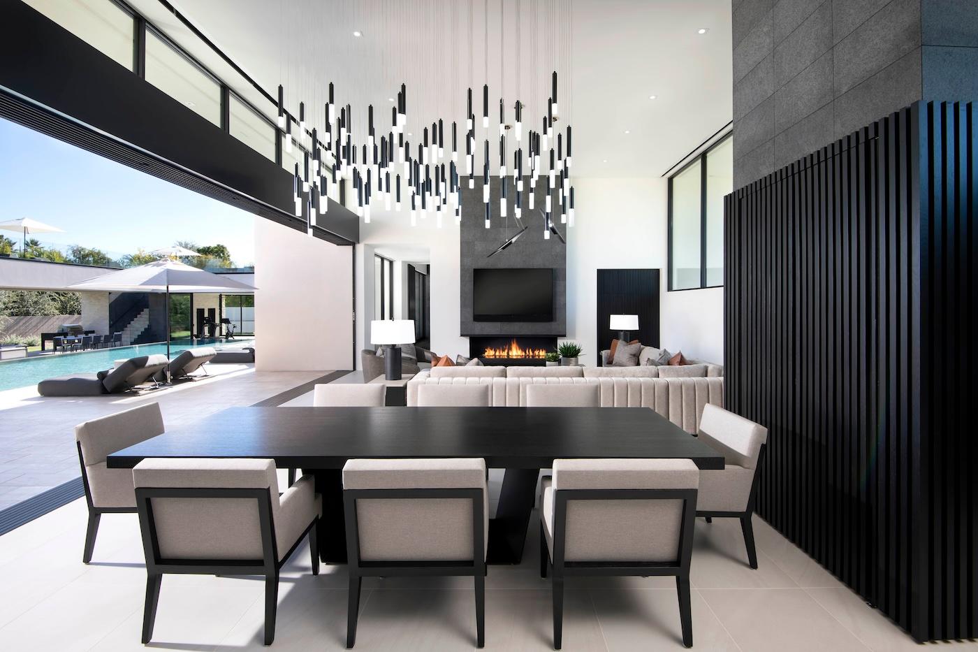 Modern living and dining area with an open layout. A black dining table is surrounded by six beige chairs. A statement chandelier hangs above. A cozy seating area by the fireplace is visible, with a view of a pool outside. The atmosphere is sleek and sophisticated.