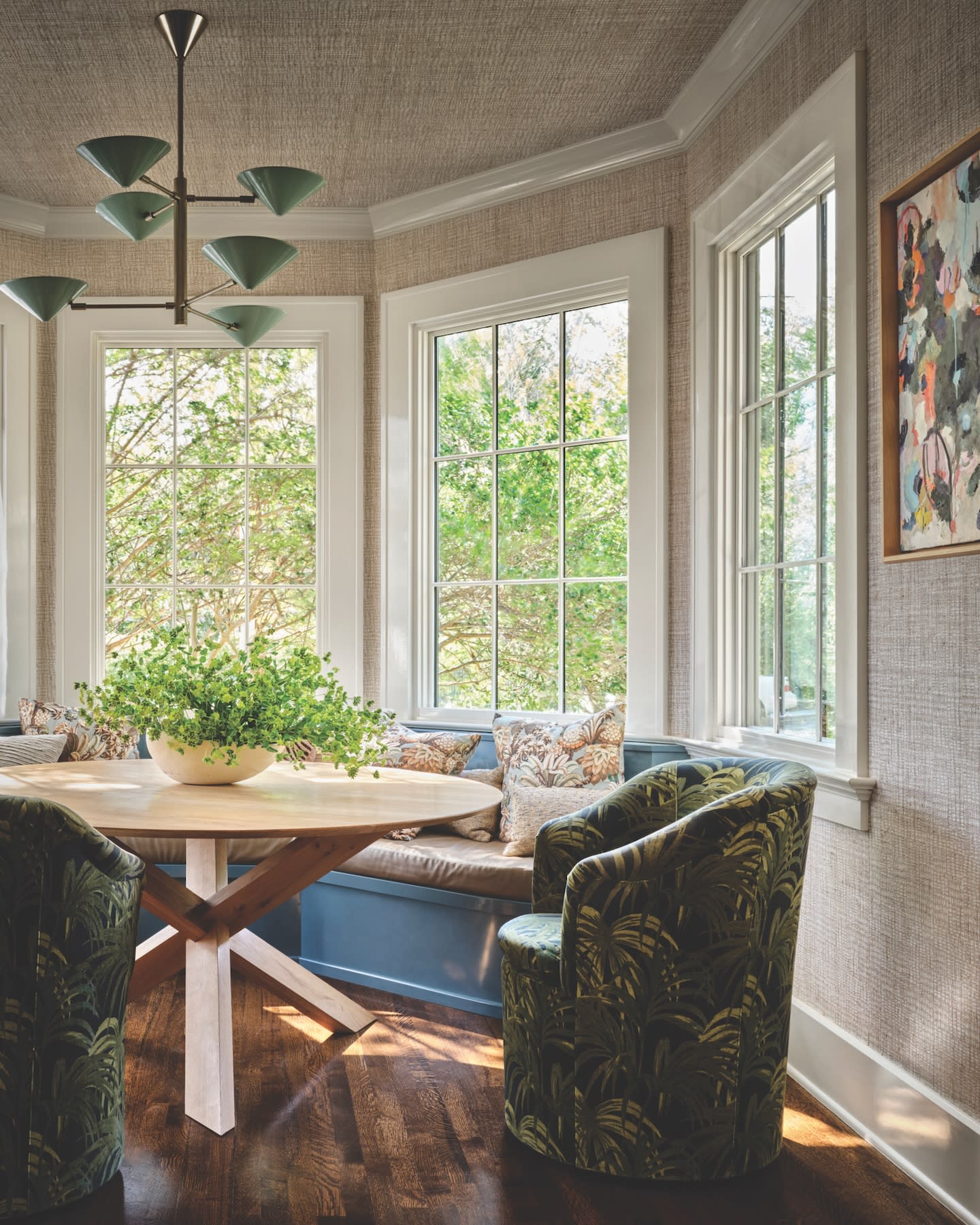 Cozy room with large windows showing greenery outside. Round wooden table with a plant centerpiece, patterned cushions on a bench, and leafy armchairs.