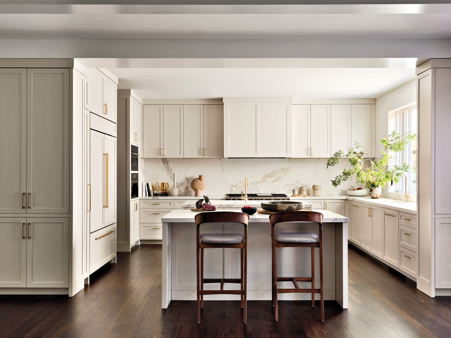 Modern kitchen with beige cabinets, marble backsplash, and dark wood floor. Central island with two wooden chairs, plants by a sunlit window. Elegant and serene atmosphere.