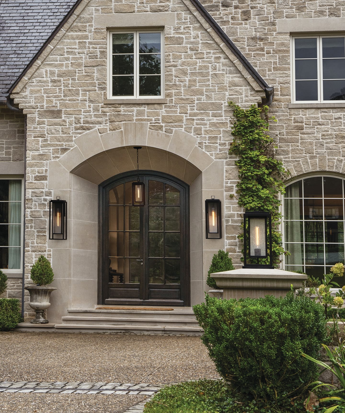 Stone facade house with arched double doors, flanked by lanterns. Ivy climbs the wall, bushes frame the entrance, creating a welcoming feel.
