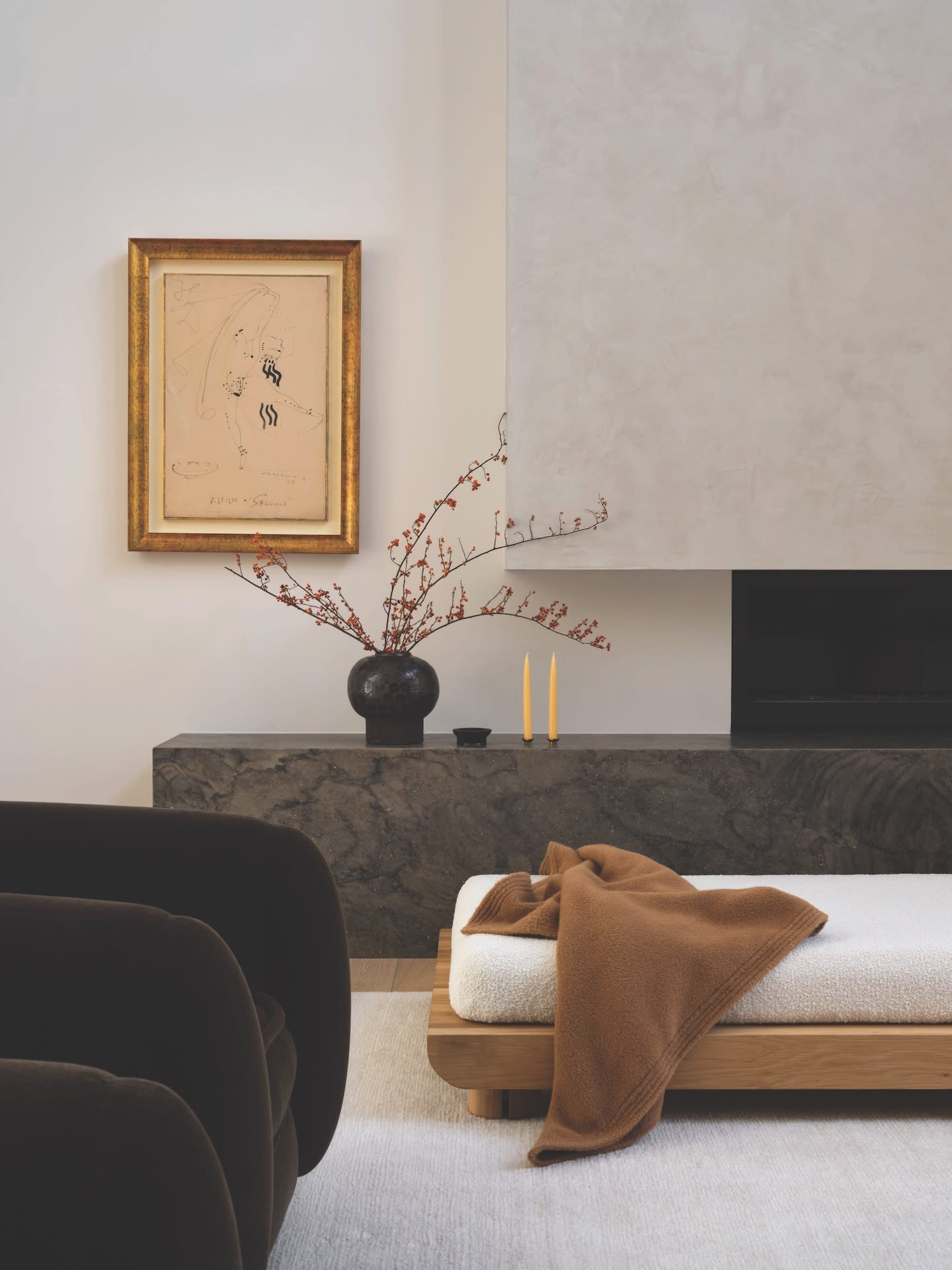 A minimalist living room with a plush, cream chaise lounge accented by a brown throw. A dark stone console holds a vase with red branches and two yellow candles, set against a neutral wall with a framed abstract artwork.