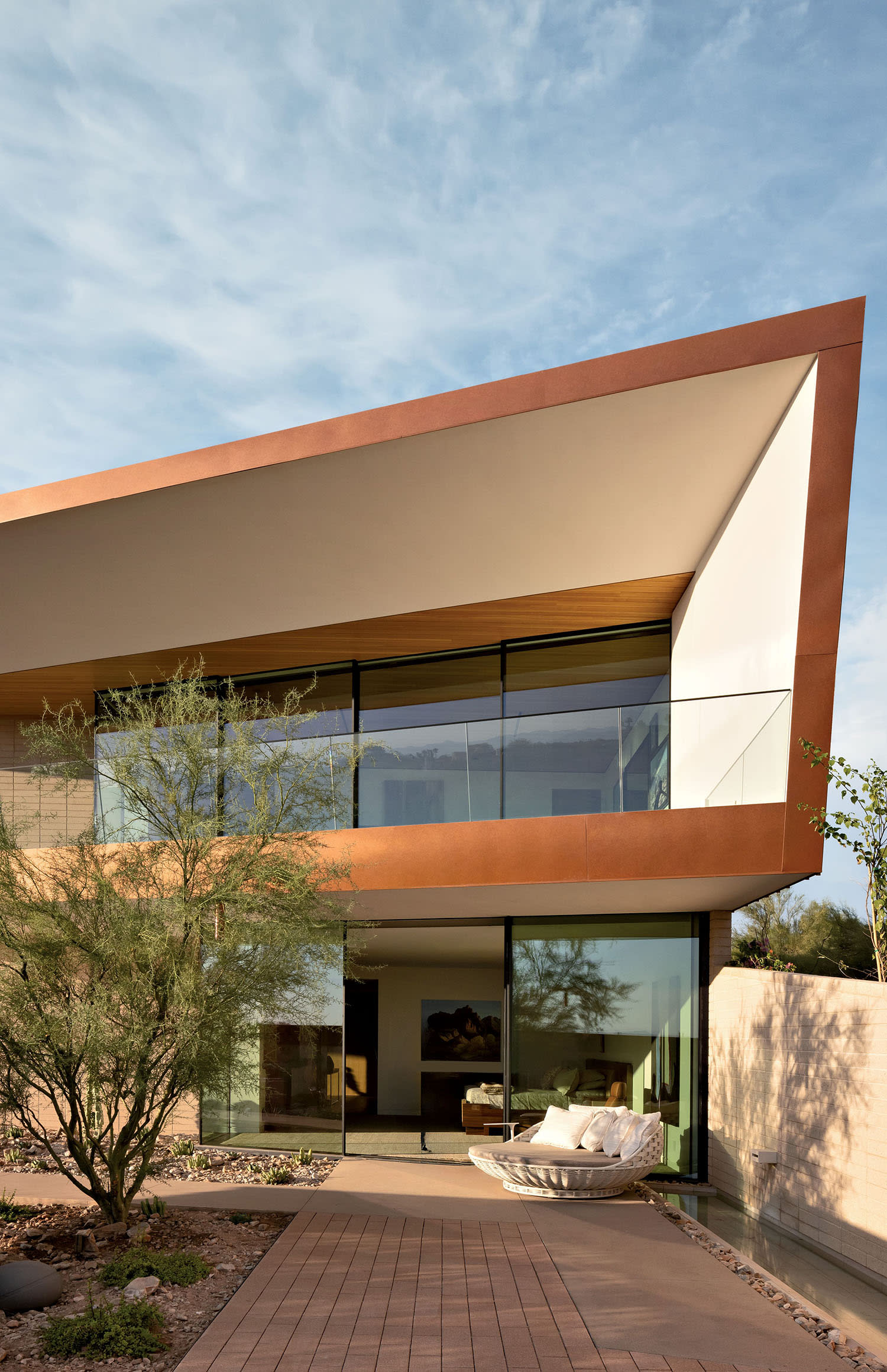 Modern house with angular roof, large glass windows, and rust-colored trim. A tree and cobblestone path lead to the entrance. Clear sky enhances tranquility.