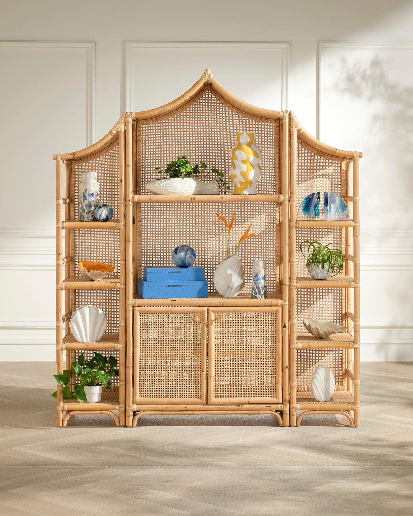 Rattan decorative shelf displaying colorful vases, plants, and sculptures against a sunlit white wall. Modern and stylish with a tropical vibe.