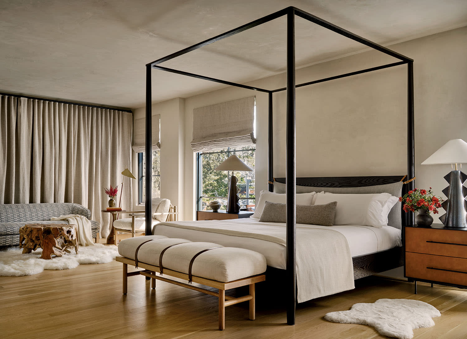 Sophisticated bedroom with a black canopy bed, beige bedding, and a plush bench. Warm lighting, large windows with nature view, and furry rugs enhance coziness.