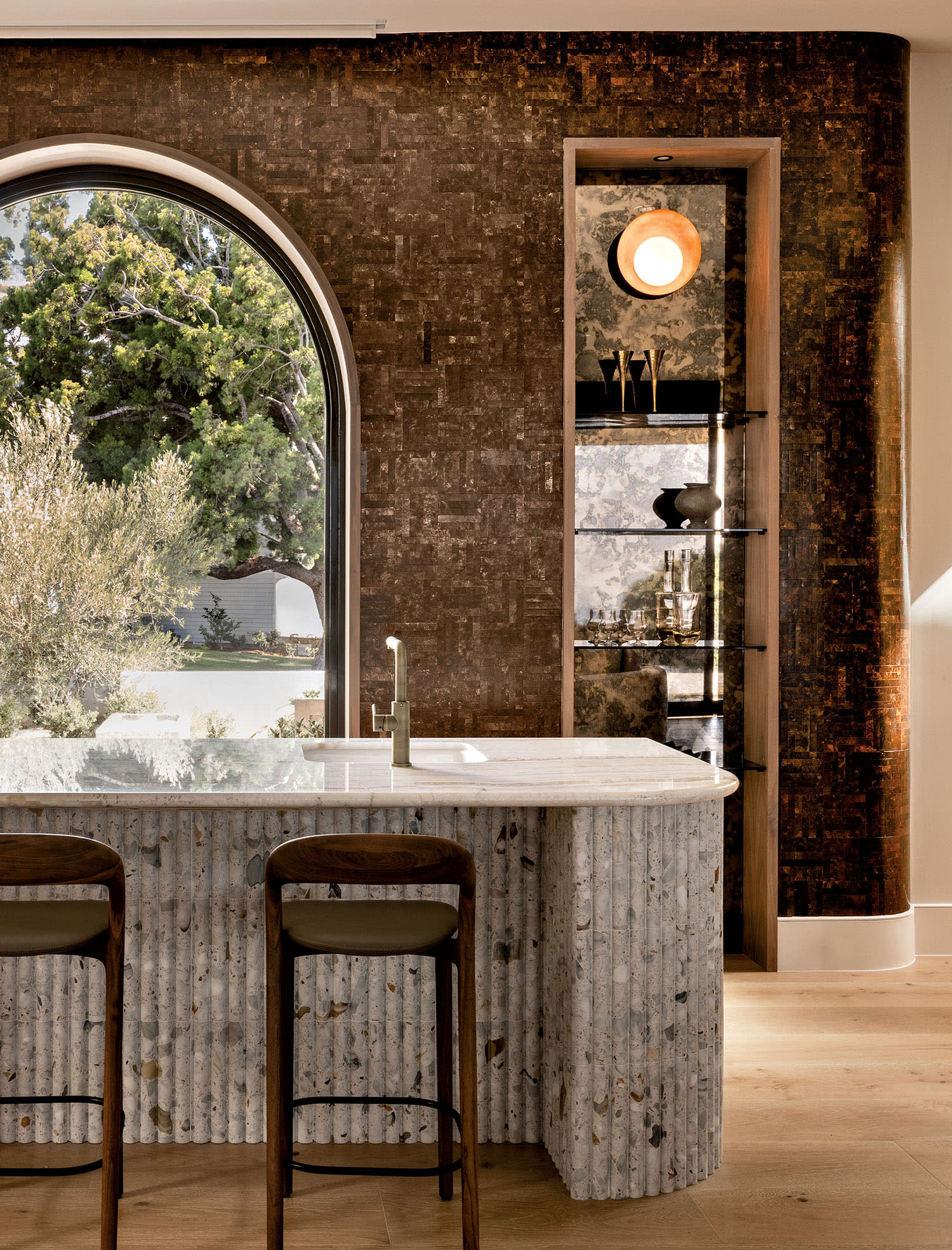 A wall of copper tile sits behind a white kitchen island with a sink and nearby high-top chairs.