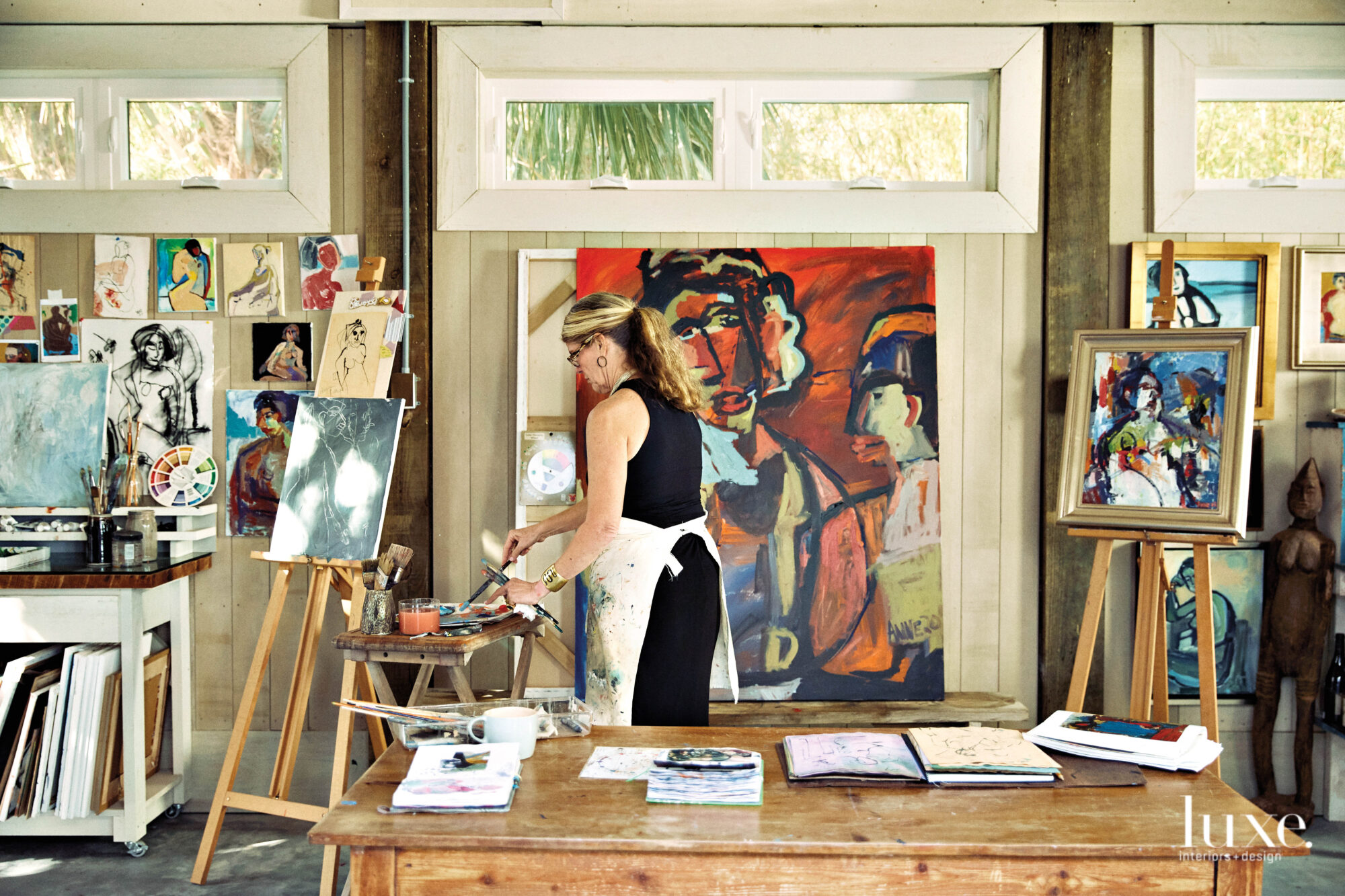 Woman standing and painting, easel with artworks to left and table in foreground