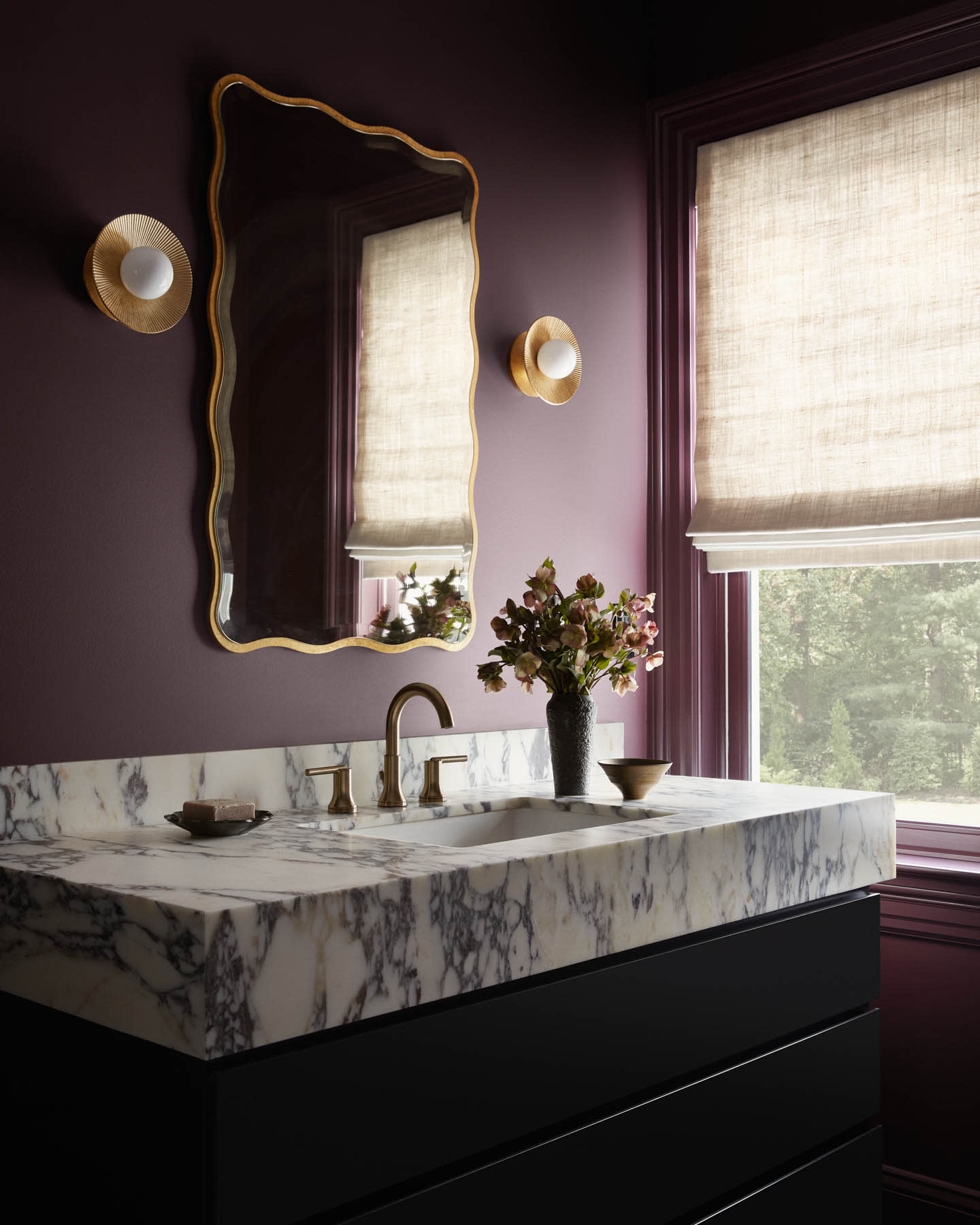 Elegant bathroom with dark plum walls and a large, ornate gold-framed mirror. A marble countertop with a brass faucet and a vase of flowers adds a touch of luxury.