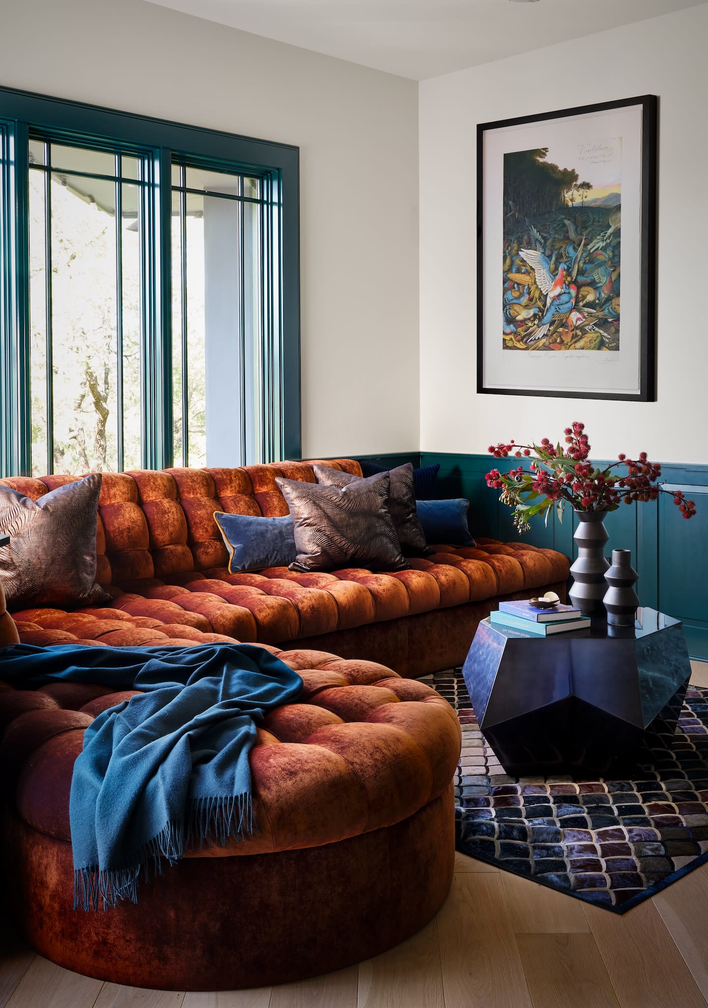 Stylish living room with a tufted, burnt orange sofa, blue throw, and geometric coffee table. Art on wall, natural light, warm and inviting.