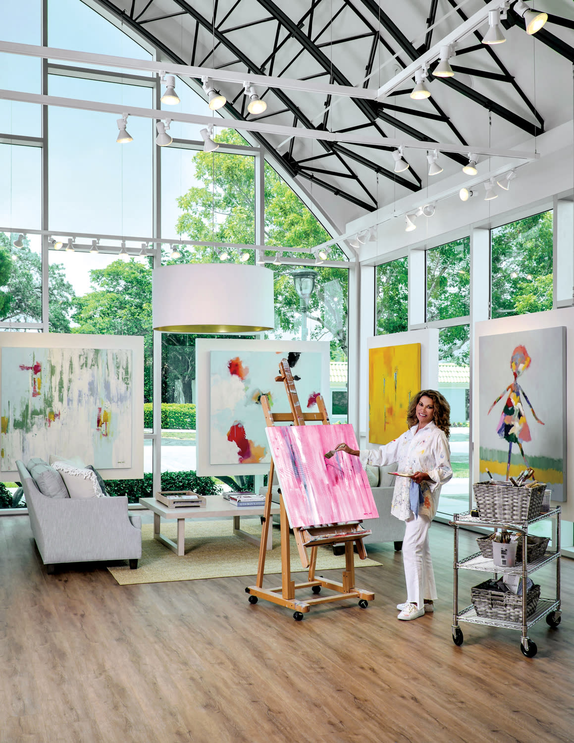 Artist Amy Moglia Heuerman paints a pink artwork in her vaulted-ceiling studio with floor-to-ceiling windows surrounded by other works.