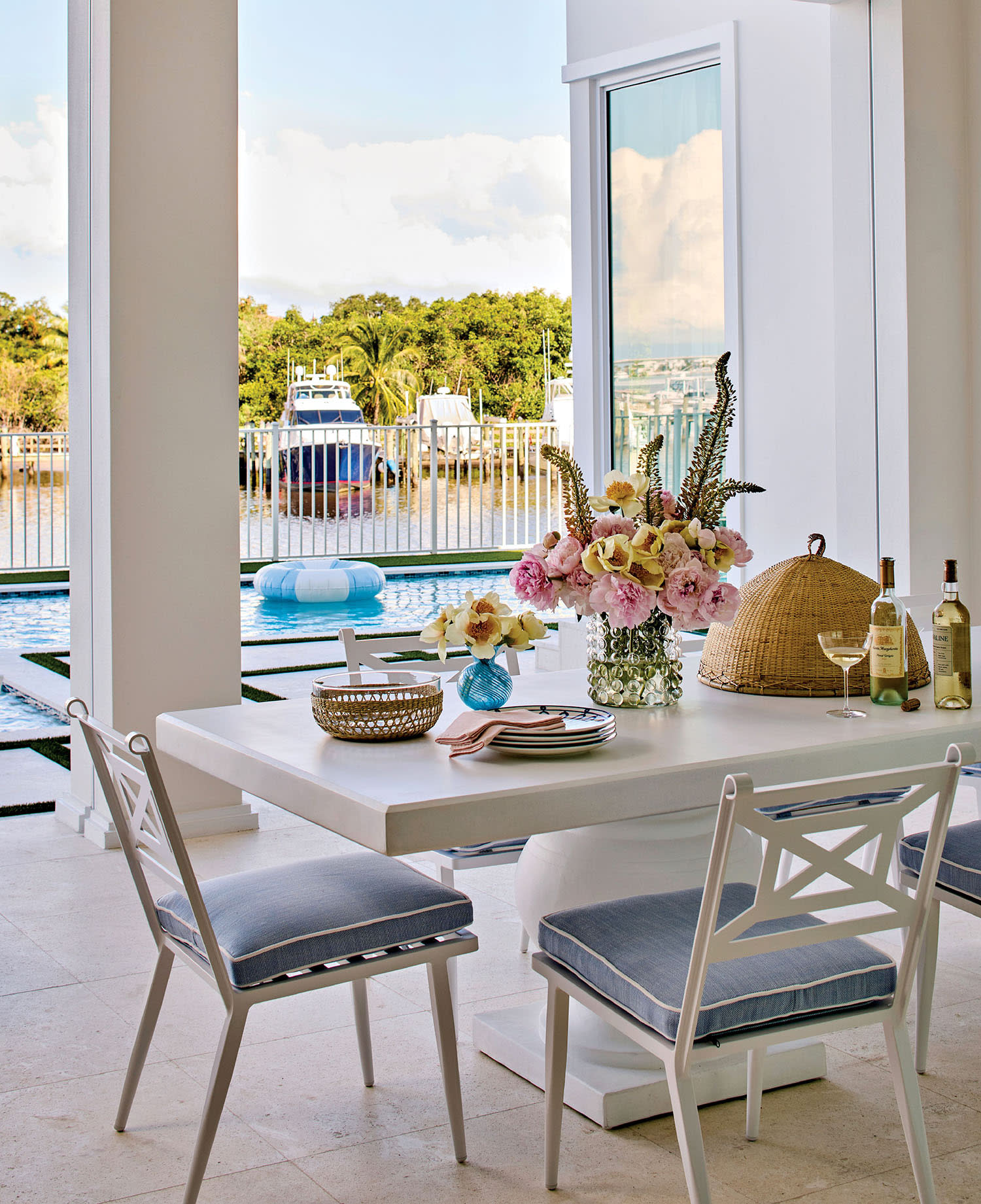 Elegant dining area with a white table, blue-cushioned chairs, and floral centerpiece. Open to a poolside view featuring a boat and lush greenery.