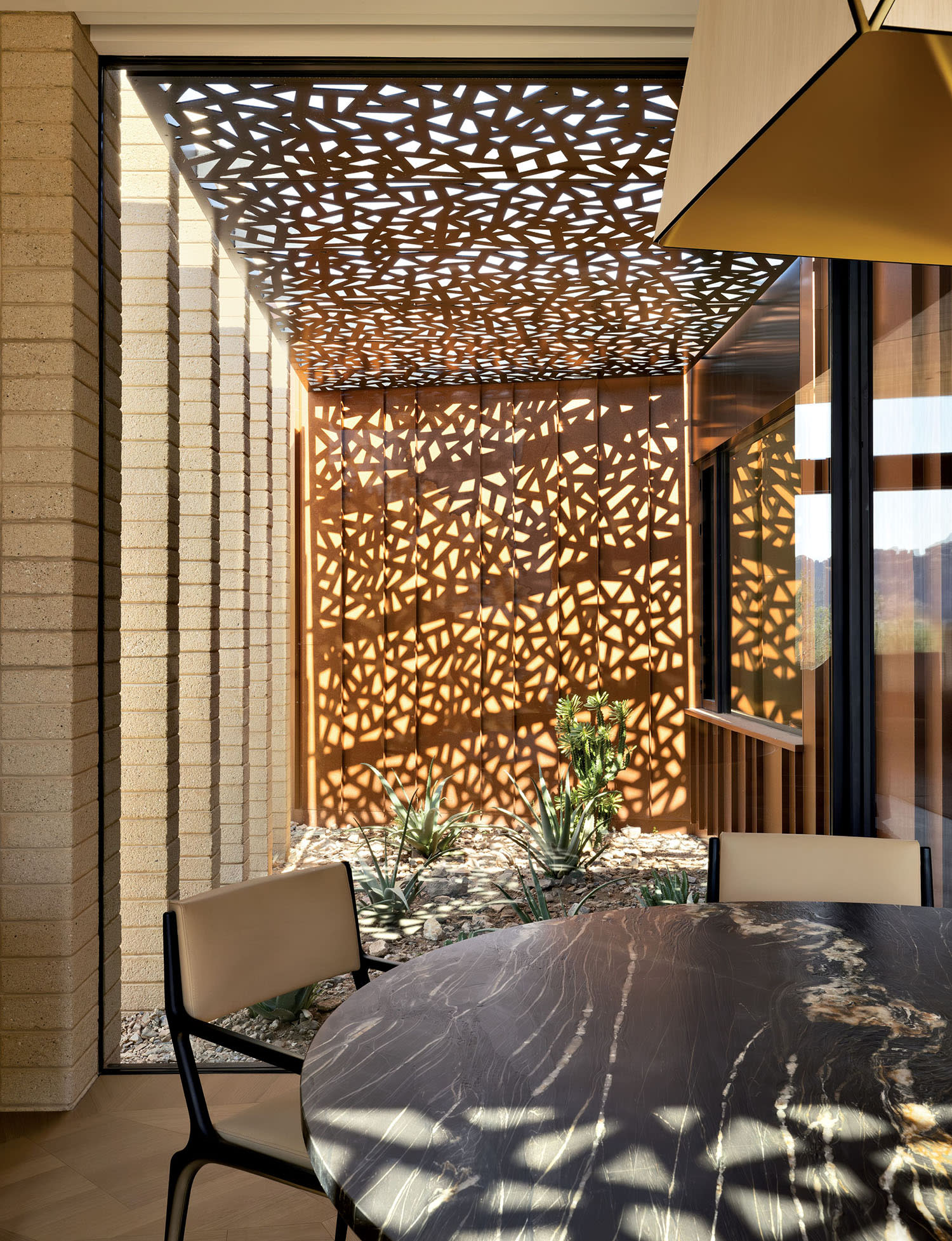 A modern interior features a large window framing a sunlit patio with geometric metal sunshades casting intricate patterns, creating a warm, artistic ambiance.