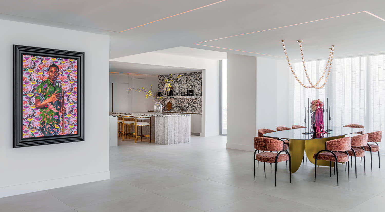 Modern interior with minimalistic design, featuring a vibrant framed artwork on a white wall. Dining area has a sleek table and pink chairs, with a chandelier above.