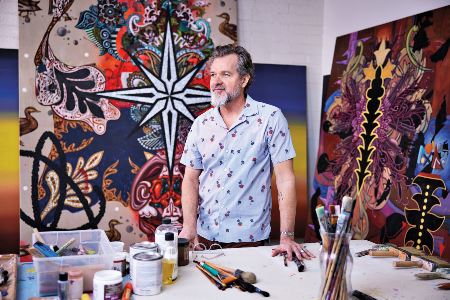 An artist stands at a table in front of two large colorful paintings.