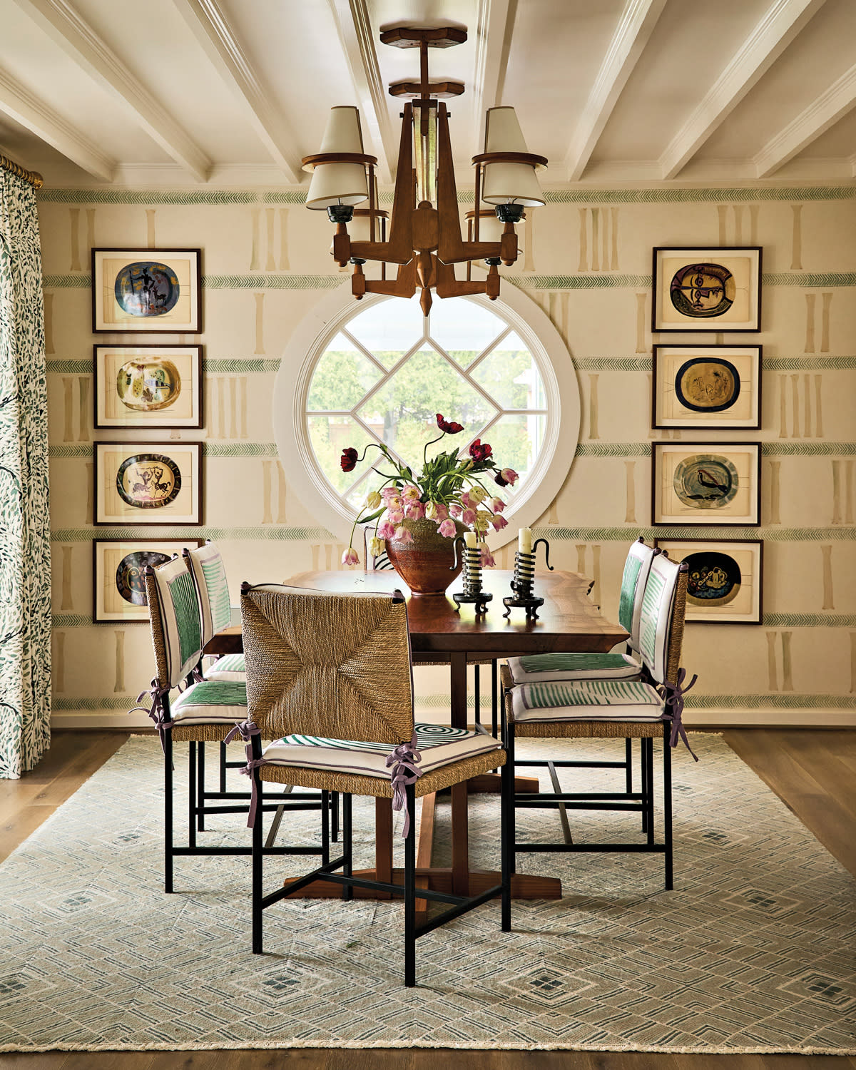 Elegant dining room with a wooden table and six wicker chairs, set against a beige wall featuring circular artwork. A round window and ceiling chandelier add charm.