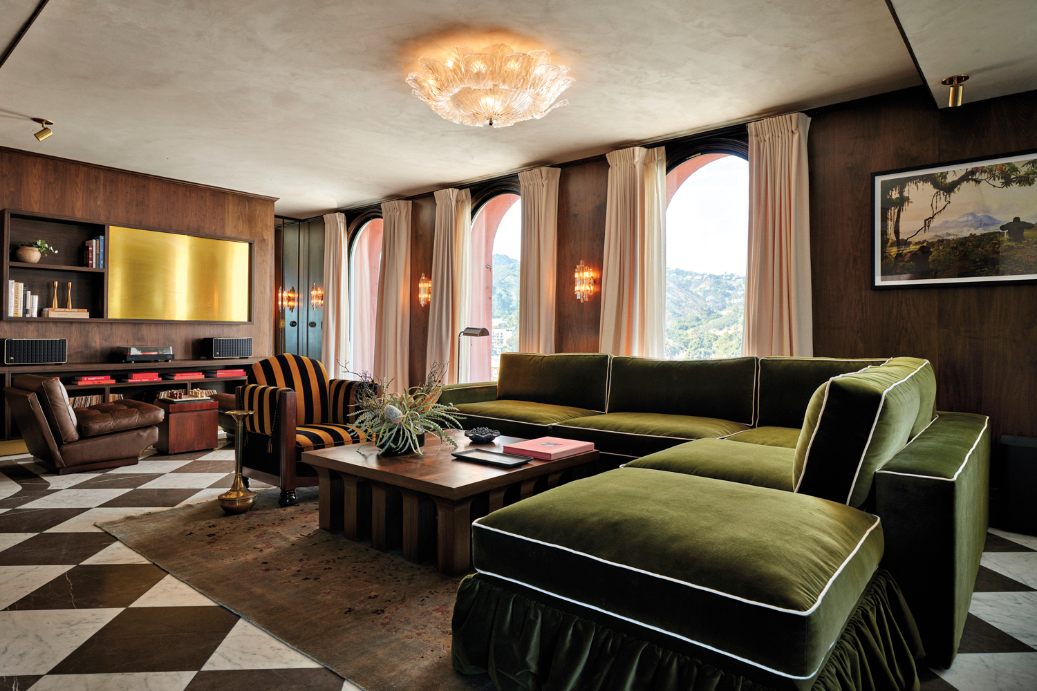 A hotel suite with marble checkerboard flooring, a green-velvet sectional, dark walls and Hollywood hill views.