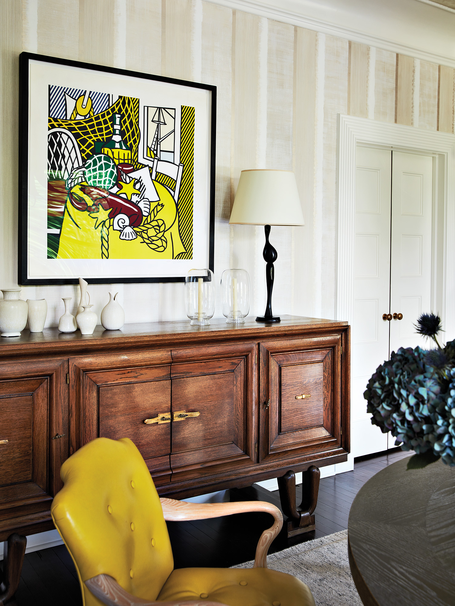 Stylish interior with a wooden sideboard, abstract art piece on the wall, and a yellow chair. A lamp on the sideboard adds a cozy touch.