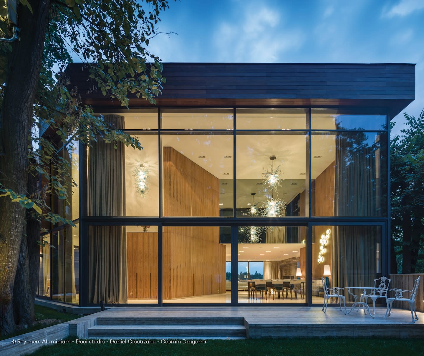 Modern glass building at dusk, with warm interior lighting. Large windows reflect the blue evening sky, surrounded by trees and outdoor seating.