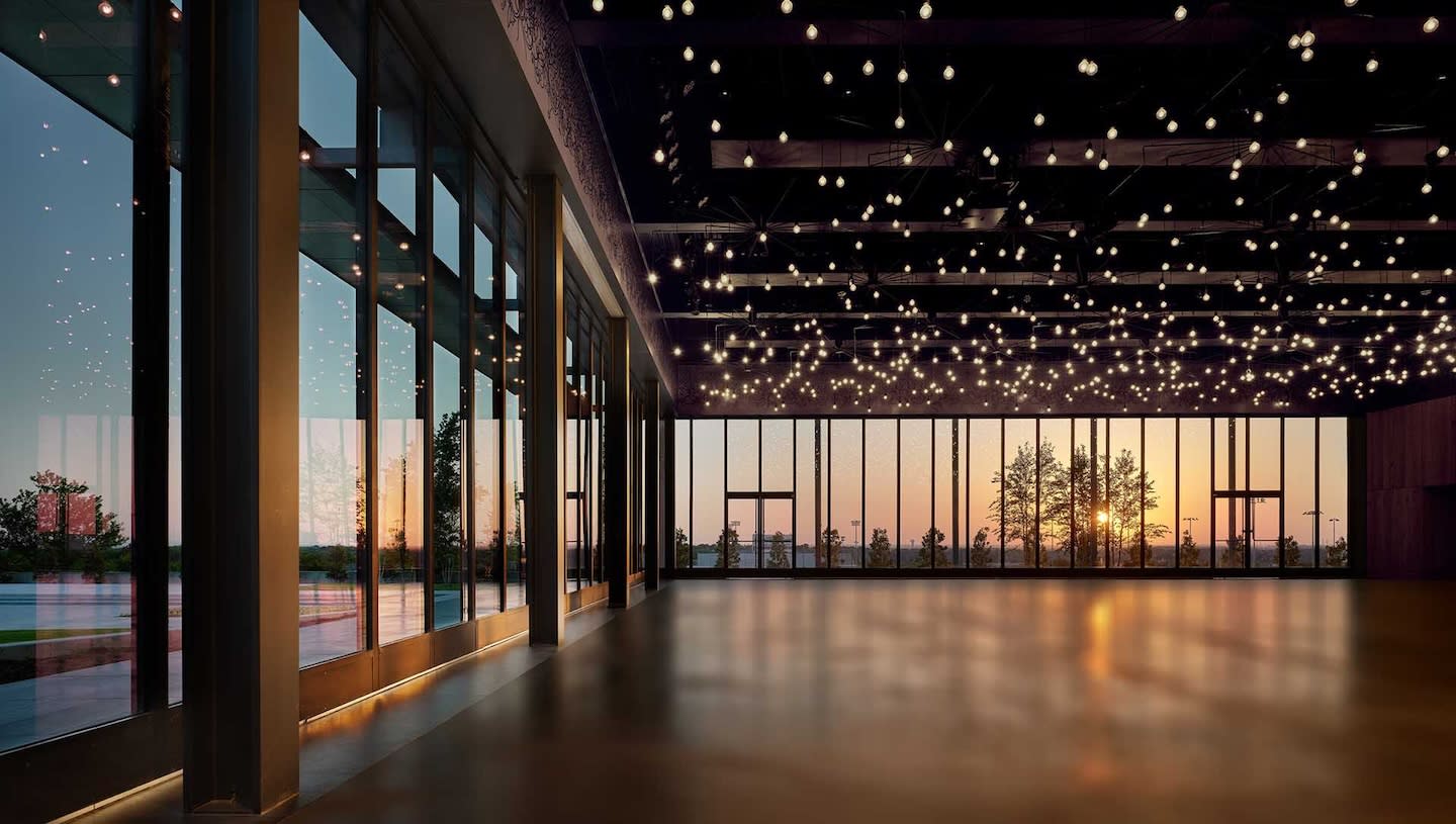 Large empty room with floor-to-ceiling windows showing a sunset view. The ceiling is adorned with numerous small, glowing lights, creating a serene ambiance.