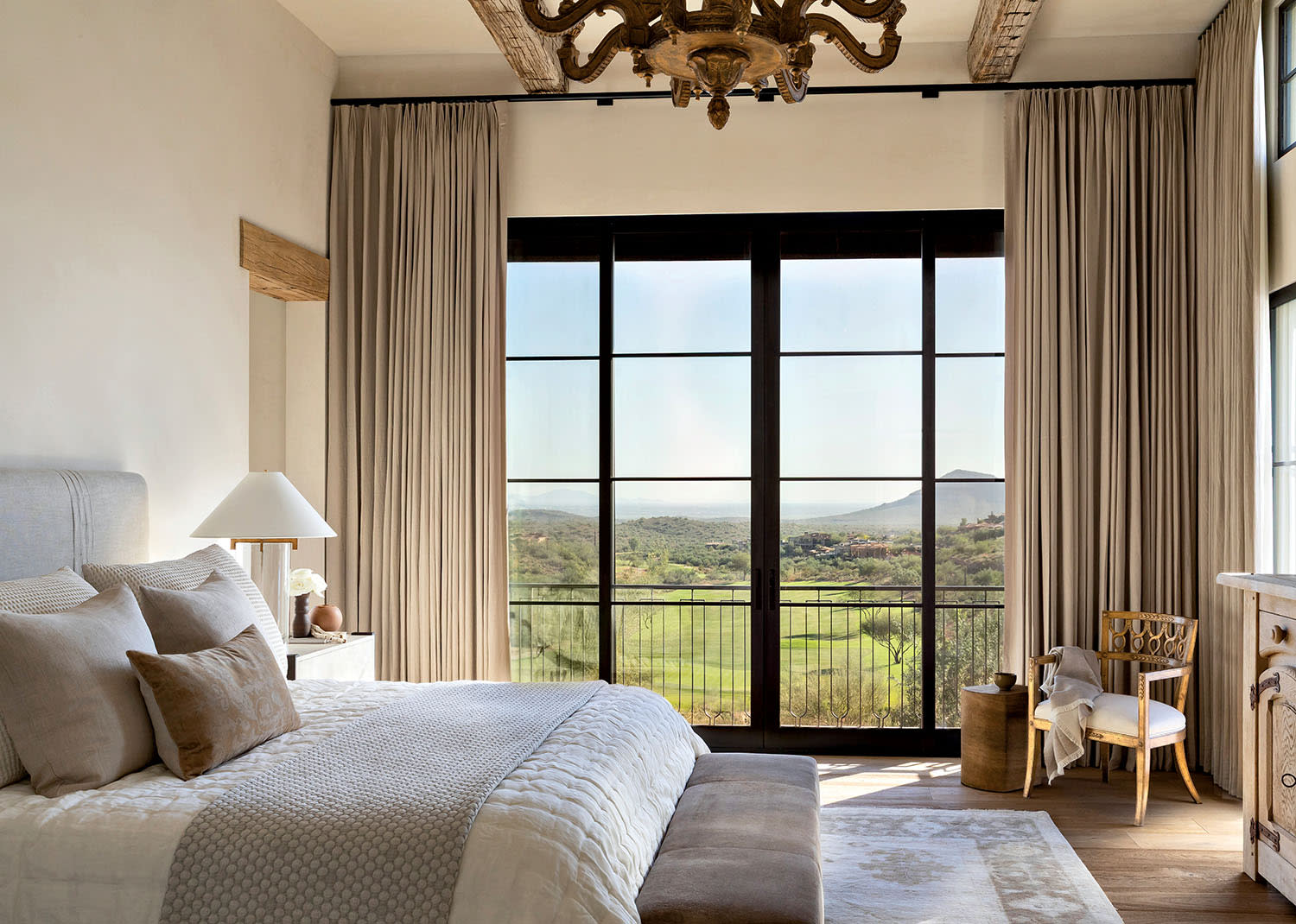 Elegant bedroom with a large window overlooking a green landscape. Neutral tones, plush bedding, and a rustic chandelier create a serene, cozy atmosphere.