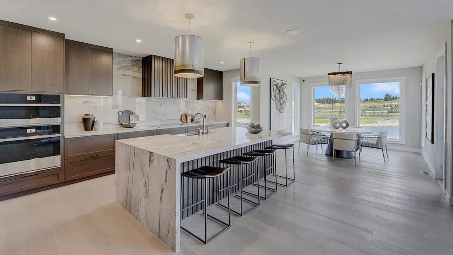 Modern kitchen with marble island, barstools, and pendant lights, leading to a bright dining area with large windows showcasing a scenic view. Elegant and spacious.