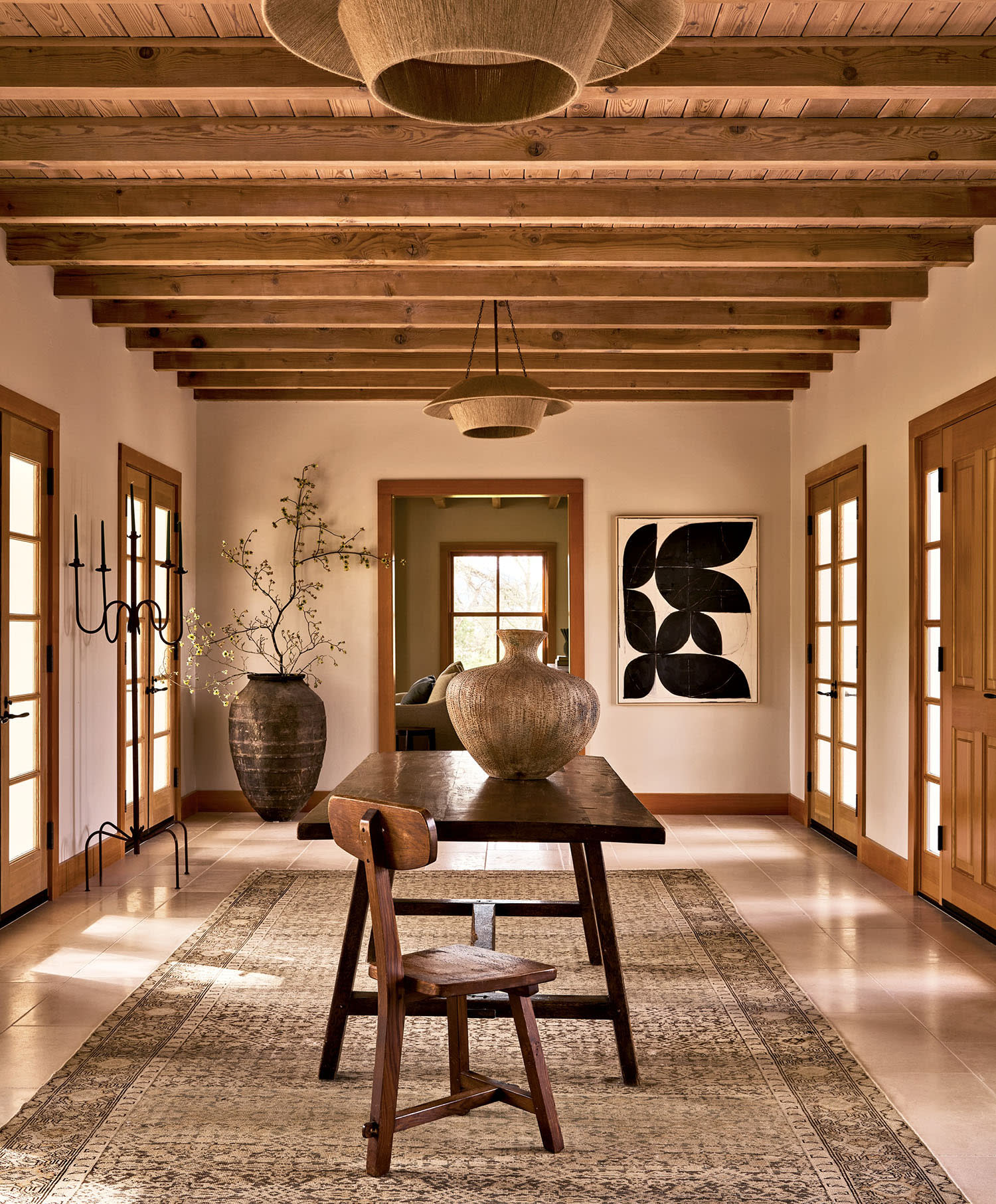 Warm, rustic hallway with wooden beamed ceiling and doors. Central table has large pottery vase. Abstract wall art and large rug enhance coziness.