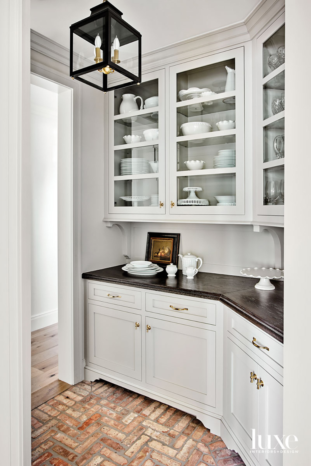 A white pantry has white cabinetry, glass-door shelves showcasing dishes and brick floors.