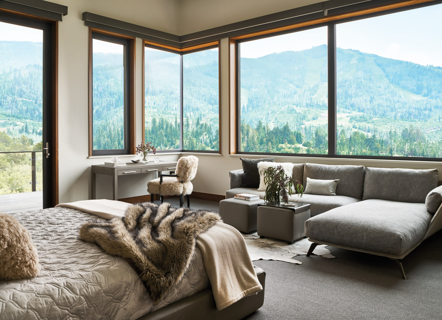 A bedroom with a bed and sectional in shades of gray and window walls facing mountain views.