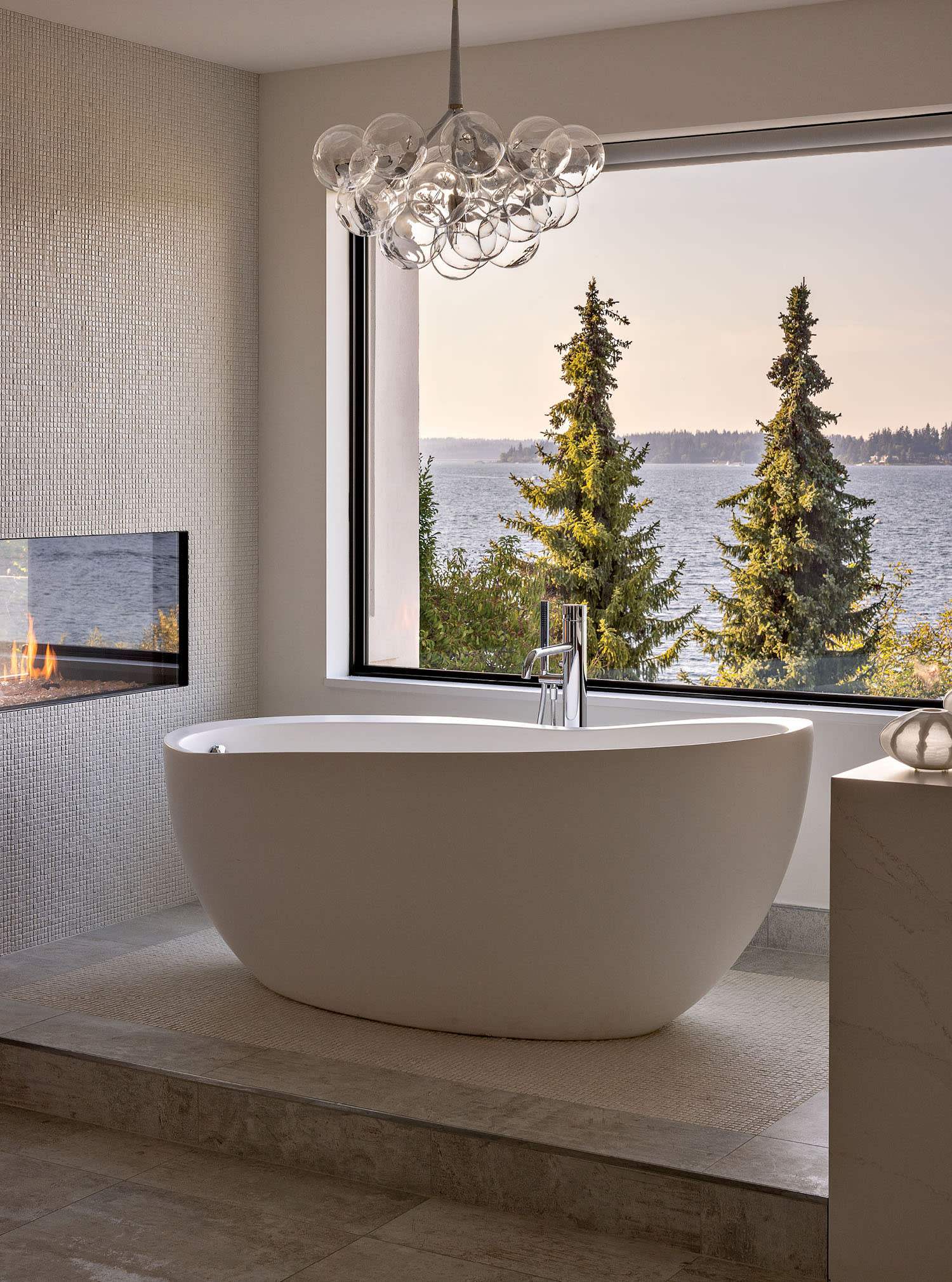 A bathtub sits on a raised platform in front of a window looking out at trees and mountains.