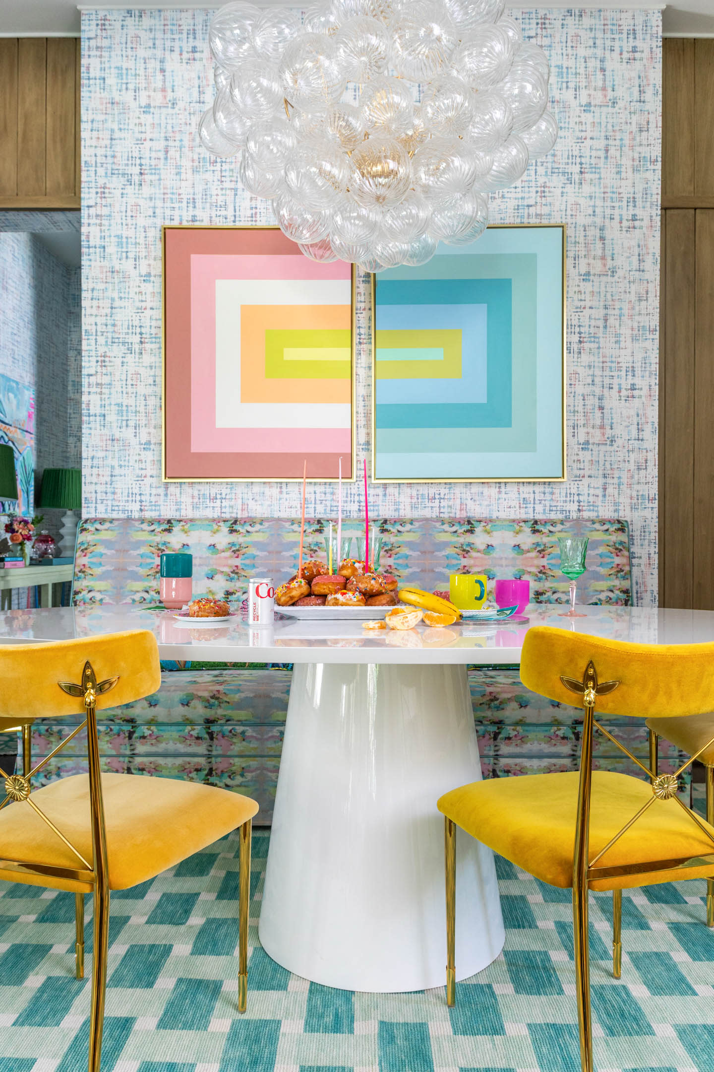 A vibrant dining area with a white table and yellow chairs on a teal-patterned rug. Bold geometric wall art, a bubble chandelier, and colorful tableware create a lively and whimsical atmosphere.