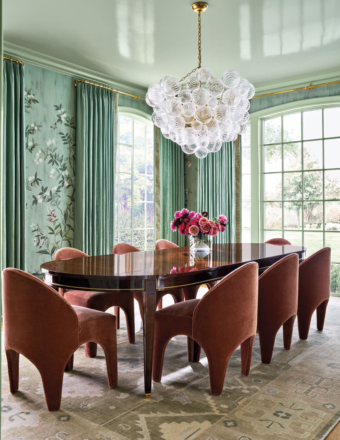 Elegant dining room with a polished wooden table, rust-colored velvet chairs, floral wallpaper, green drapes, and a cluster glass chandelier.