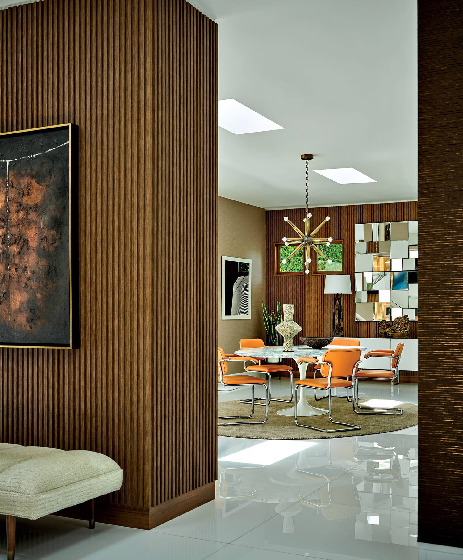 A room with wood paneled walls, modern artwork, and a geometric chandelier sitting above a table with orange chairs. 