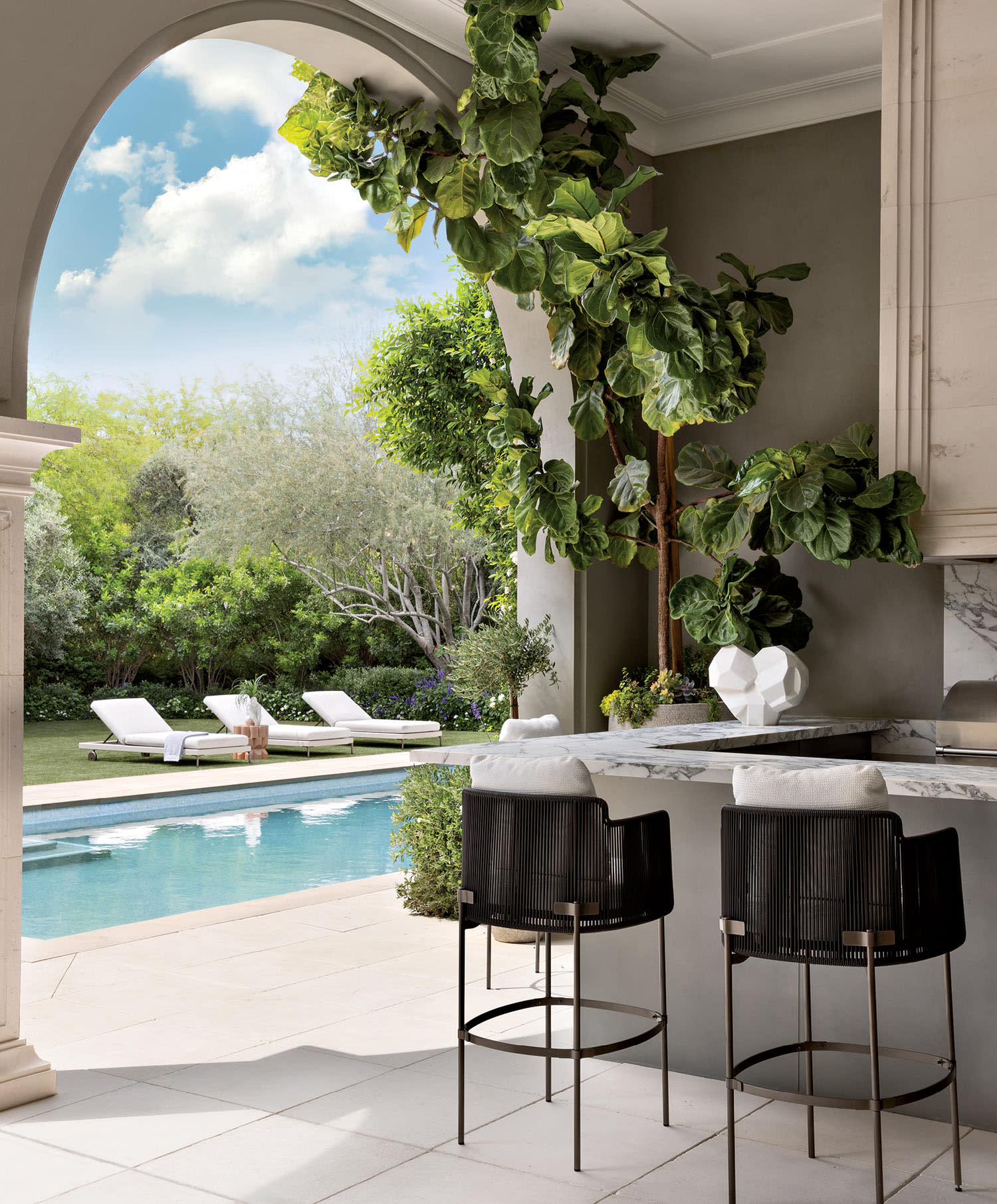Luxurious outdoor patio with a pool, two black chairs at a marble bar, and lush greenery, creating a serene, inviting atmosphere under a bright sky.