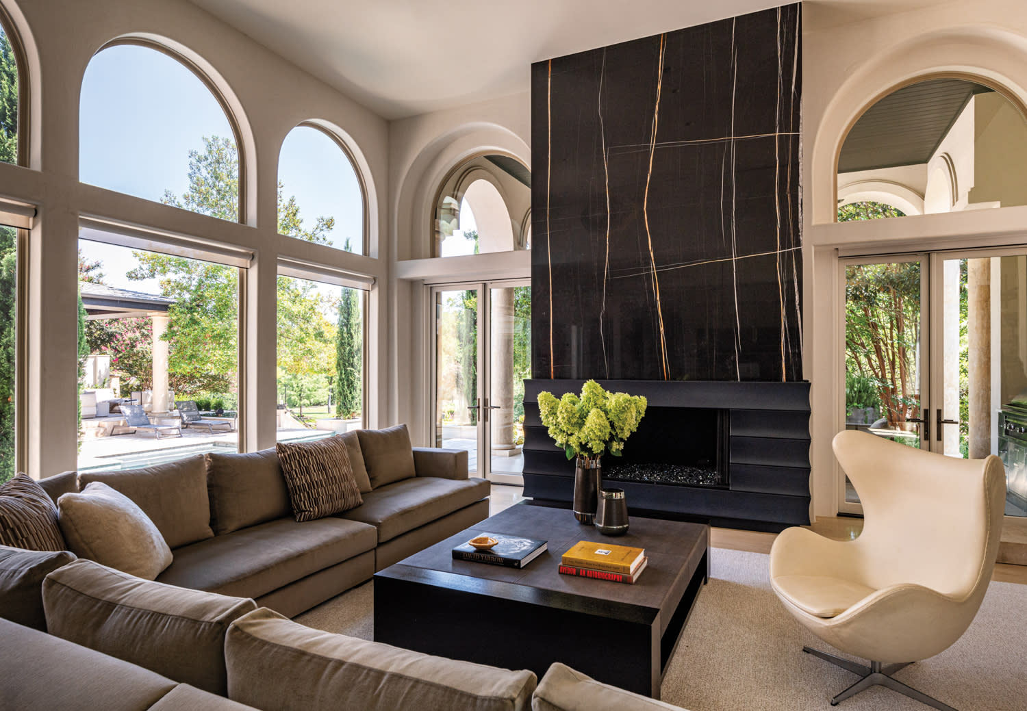 Modern living room with a high black marble fireplace, arched windows, beige couches, a large square coffee table, and a white swivel chair.