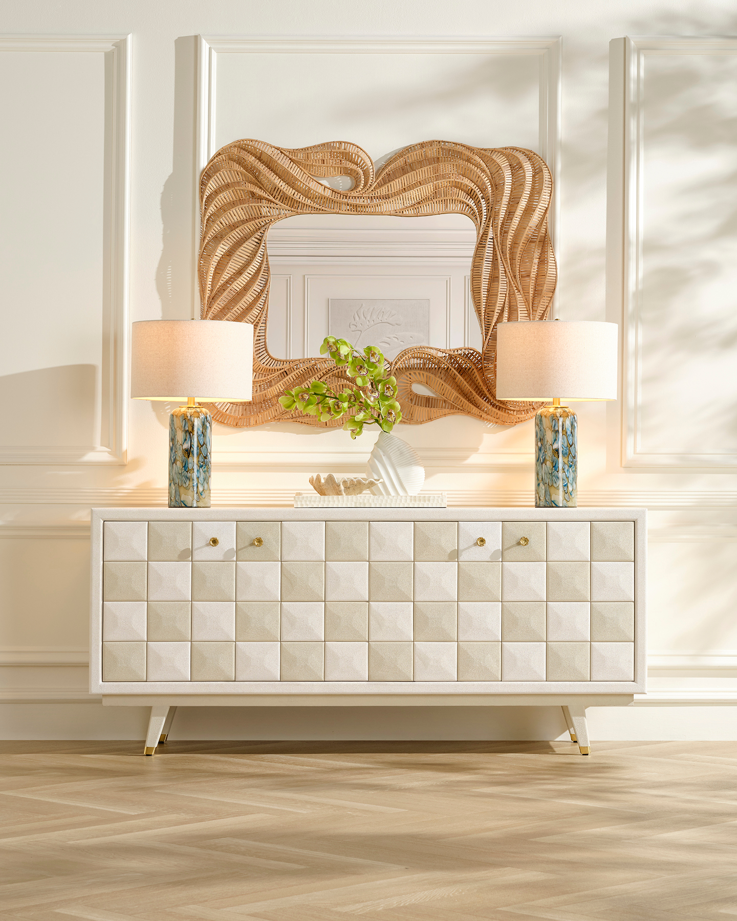 Elegant living room with a textured sideboard, flanked by two blue marble lamps with beige shades. Above, a wavy rattan mirror and leafy decor add flair.