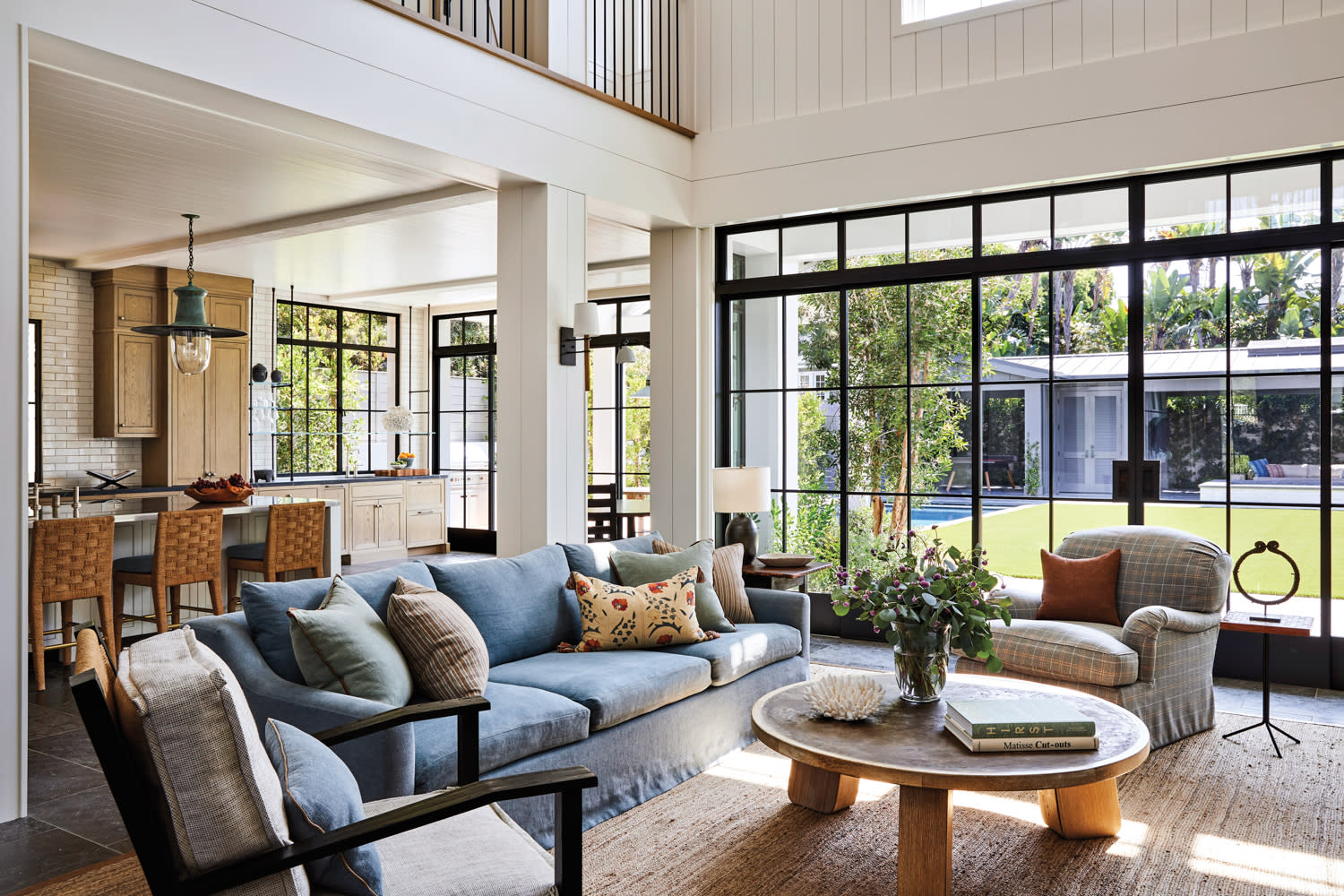 Open living room with steel-framed window wall, blue sofa, light-wood coffee table and kitchen in the back