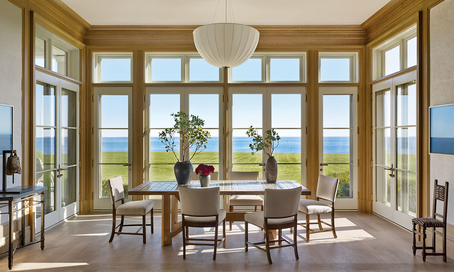 Elegant dining room with large windows overlooks the ocean. A wooden table with chairs and potted plants adds a serene, coastal ambiance.