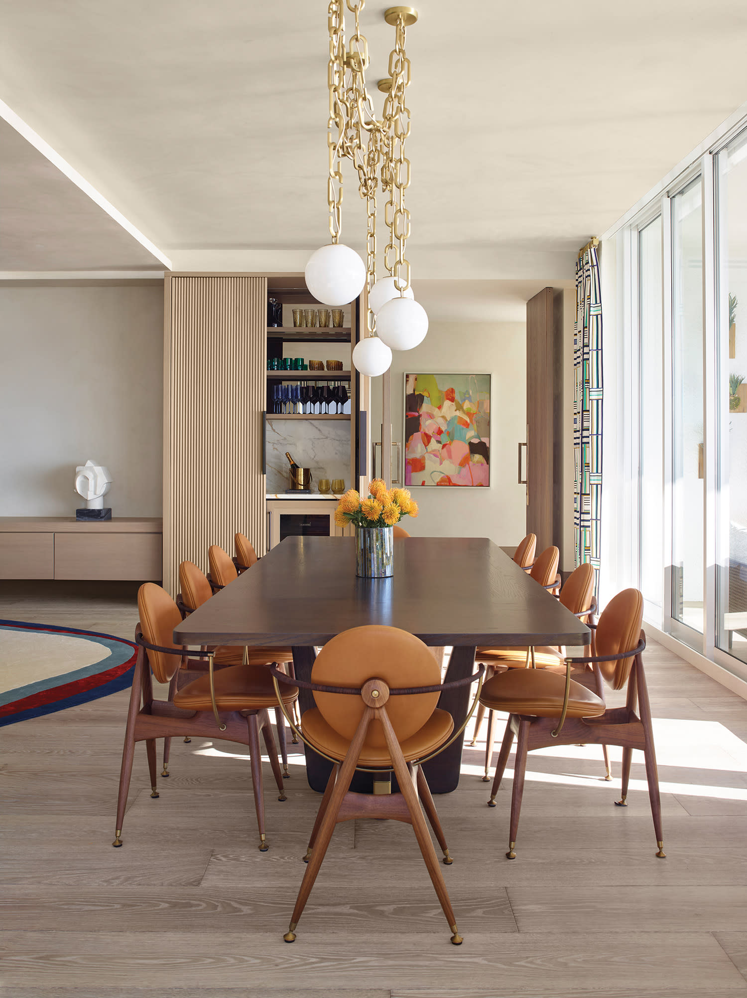 Spacious dining room with a sleek, dark wood table, surrounded by tan leather chairs, beneath a gold chandelier. Large windows and abstract art add brightness.