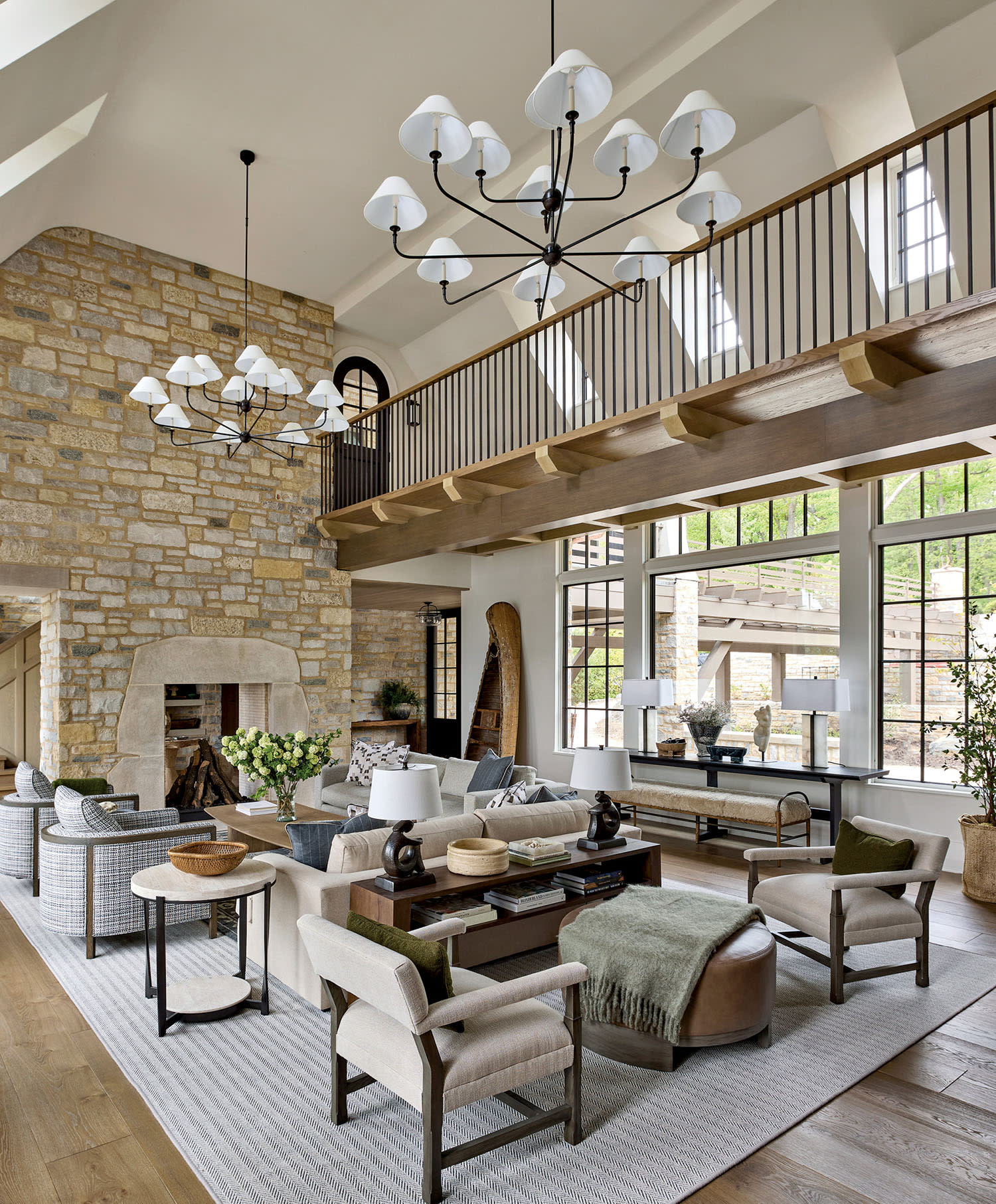 Spacious living room with a stone fireplace, wooden floors, and large windows. Light decor includes a cozy seating area and modern chandeliers, creating an inviting atmosphere.