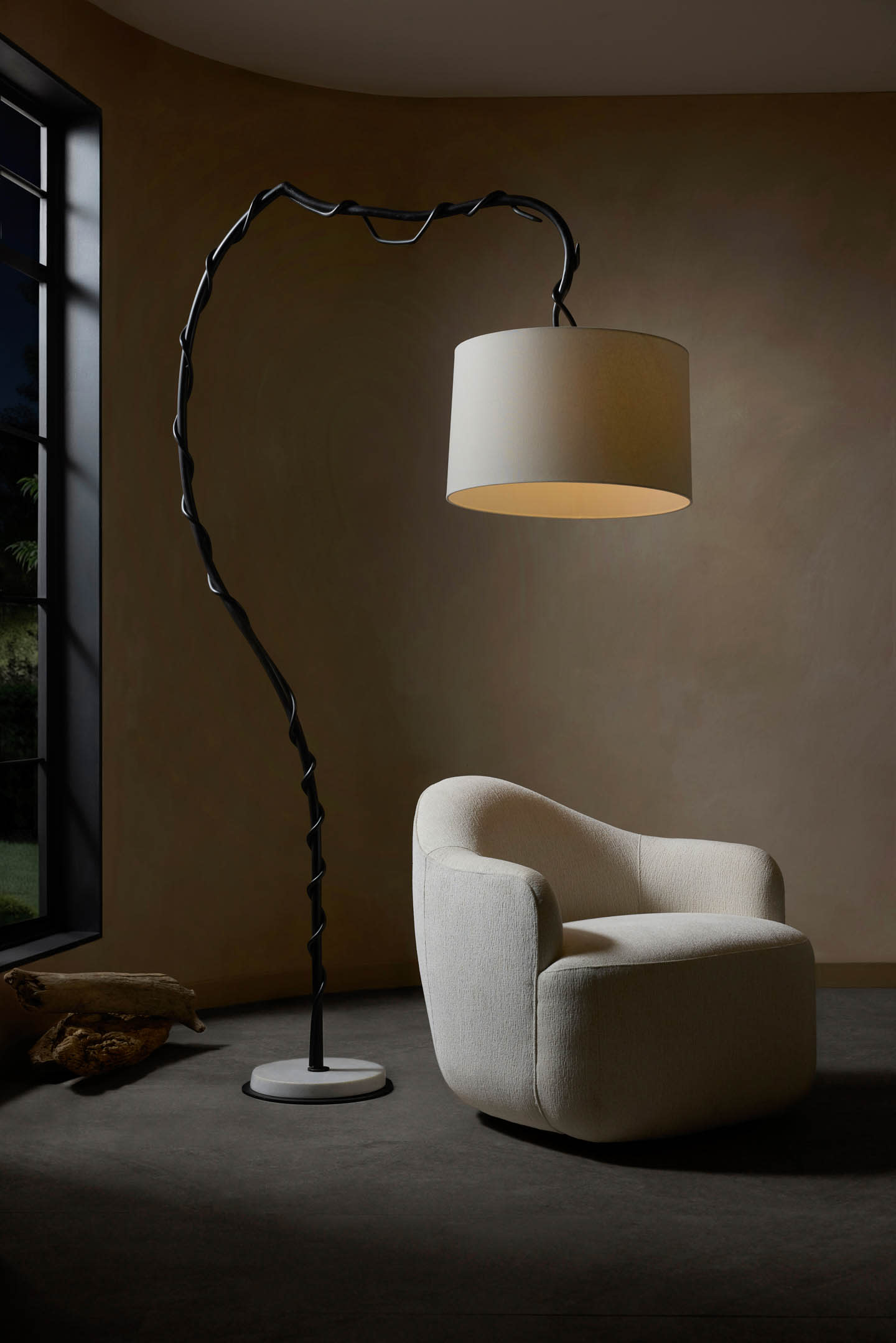 A modern, curved floor lamp with a large, round shade stands beside a plush, white armchair in a dimly lit room with minimalist decor.