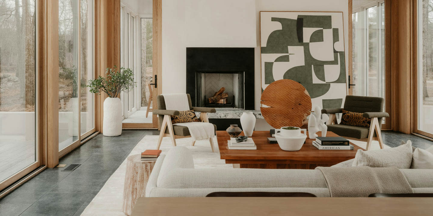 Modern living room with large windows, a central wooden table, abstract art, and a fireplace.