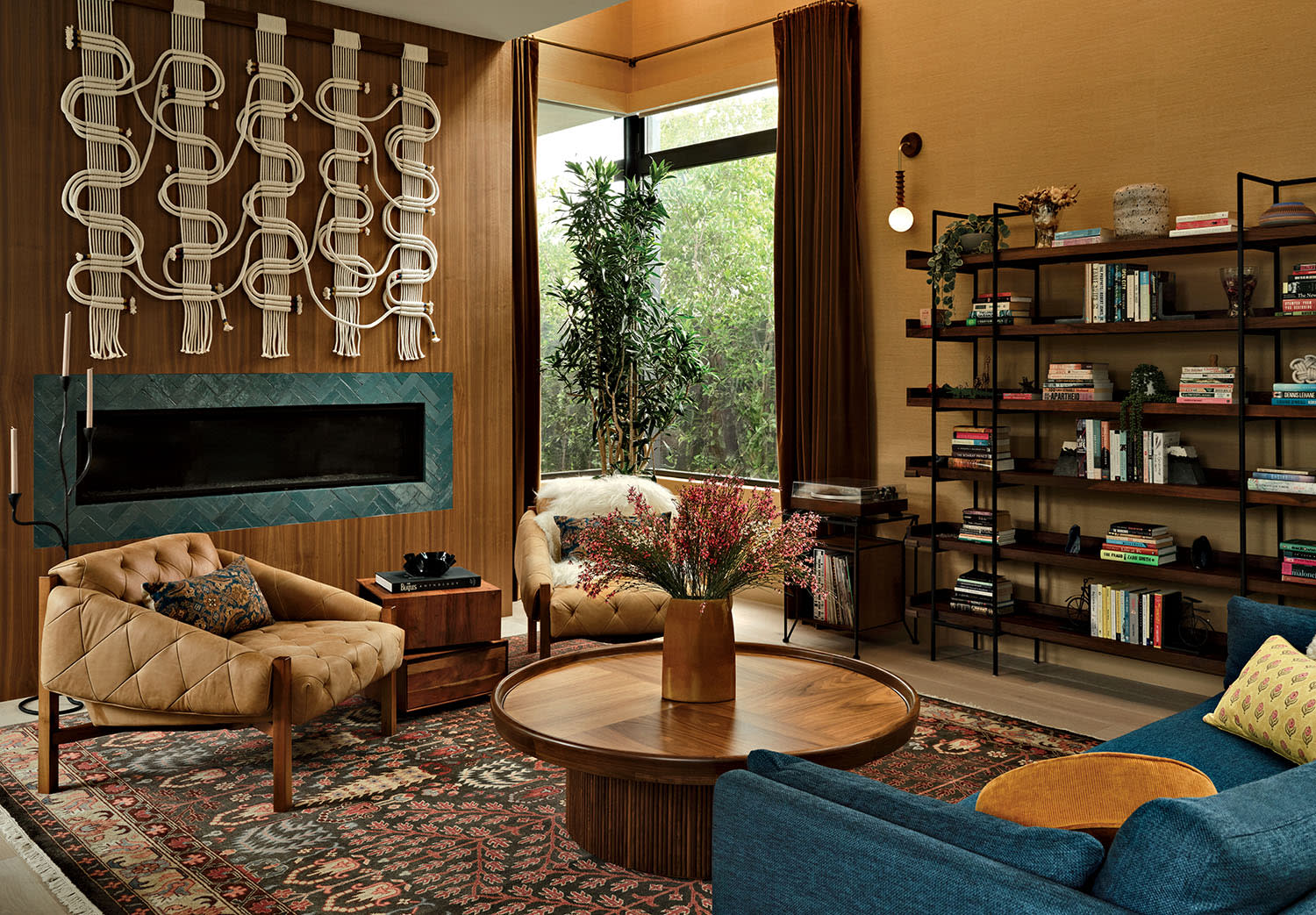 A cozy living room with warm tones features a modern fireplace, tufted chairs, a blue sofa, a round wood table, a patterned rug, and a bookshelf.
