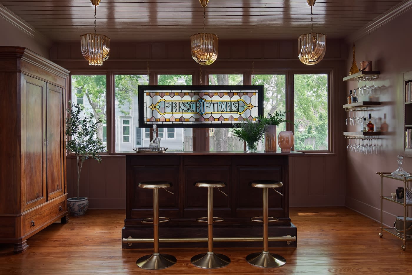 Elegant home bar with stained glass "Prescriptions" sign, wooden cabinetry, three gold stools, and chic lighting, exuding a cozy, stylish atmosphere.