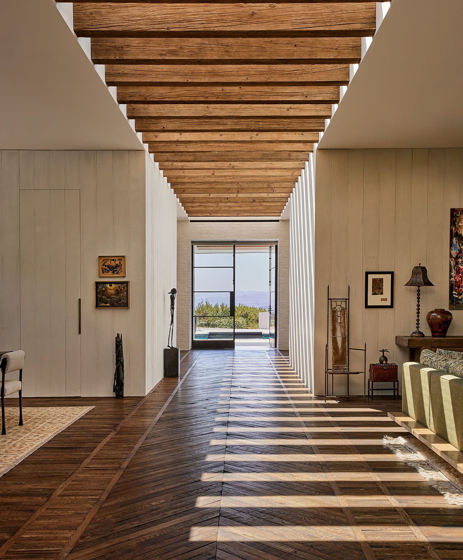 Spacious hallway with wooden ceiling beams and floor, leading to a sunlit window view. Art pieces and furniture adorn the sides, creating a warm ambiance.