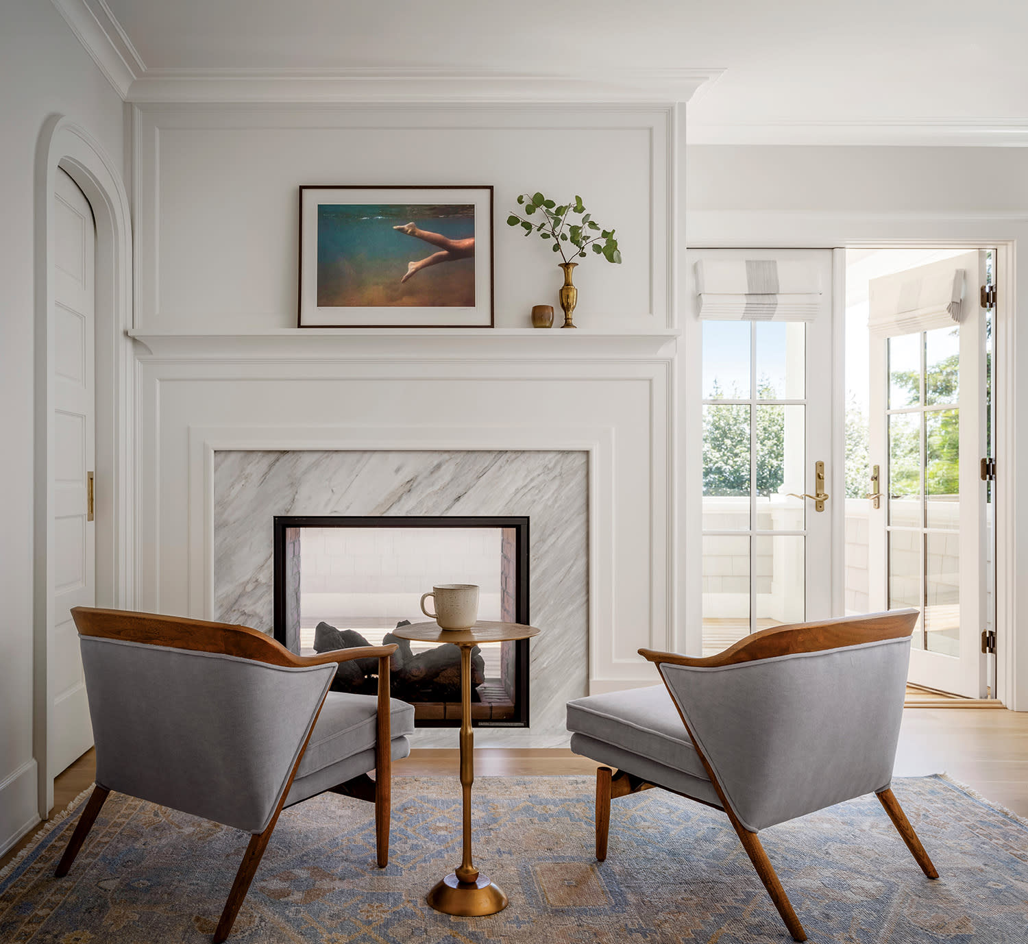 Elegant living room with two gray mid-century chairs facing a white marble fireplace. A small round table holds a mug. Sunlight streams through open French doors.