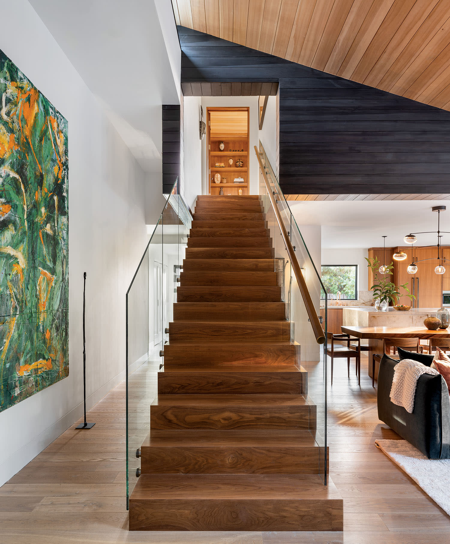 A modern wooden staircase with glass railings leads to a loft. The interior features bright lighting, a large abstract painting, and a cozy living space.